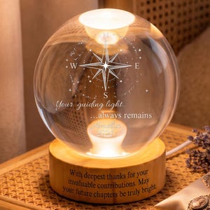 Może przedstawiać: Sferyczna szklana lampa z wygrawerowanym wzorem róży wiatrów, napisem "Your guiding light... always remains", imieniem oraz rokiem "2026". Szklana kula umieszczona jest na cylindrycznej drewnianej podstawie z wygrawerowaną długą wiadomością.