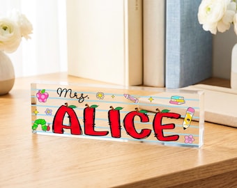 以下が含まれることがあります： Mrs. ALICEという名前が太い赤文字で書かれた、透明なアクリルの長方形デスクサイン。背景は罫線入りのノート用紙を模しており、緑色の毛虫、ピンクのリンゴ、鉛筆、本のカラフルなイラストが描かれています。
