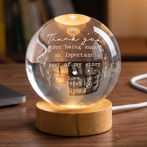 May include: A clear glass sphere with the words "Thank you" and "For being such an important part of my story" etched on the surface. The sphere sits on a wooden base and is illuminated from within, with the text "Love Sarah" below.