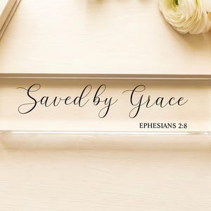 May include: A clear acrylic block with the words "Saved by Grace" in elegant black script. Below the text is "Ephesians 2:8" in a smaller, simple font. The block is rectangular and sits on a light-colored surface.