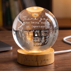 May include: A clear glass sphere lamp resting on a cylindrical light wood base. The sphere is engraved with the text: Thank you For being such an important part of my story Love Sarah, surrounded by floral designs. The wood base also says Thank you.