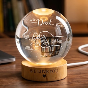 May include: A clear glass sphere with the words "Best Dad Ever" and a graphic of clasped hands, illuminated by a warm light. The sphere sits on a wooden base engraved with "We Love You" and a heart symbol. The names Olivia, Sarah, and Daddy are also visible.