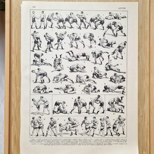 Puede incluir: Una impresión antigua que representa movimientos de lucha libre, presentada en una bandeja de madera. Una lupa dorada y un bolígrafo color crema también están en la bandeja. Una etiqueta con un logotipo circular está adjunta a la impresión.