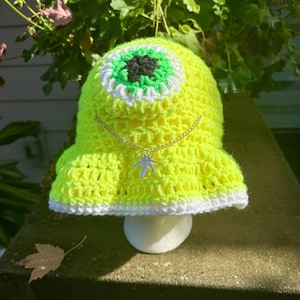 Peut inclure: Un chapeau au crochet jaune néon avec un grand motif d'œil au centre, avec des détails blancs, verts et noirs. Une chaîne argentée avec un charme en forme de feuille est suspendue au chapeau. Le chapeau a un bord blanc.