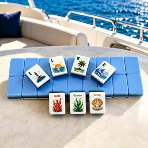 Puede incluir: Un juego de fichas de Mahjong sobre una base azul claro. Las fichas presentan varias ilustraciones, como un faro, una puesta de sol, una palmera y coral. Las fichas est&aacute;n dispuestas sobre una superficie blanca con un yate al fondo.