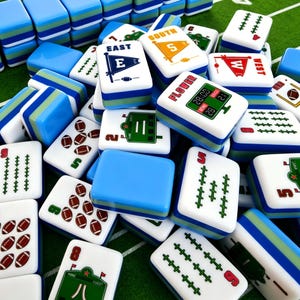 May include: A collection of white mahjong tiles featuring football-themed graphics, including directional flags labeled East, South, and West, football icons, and scoreboard designs. The tiles have blue and green layered bases and are scattered.