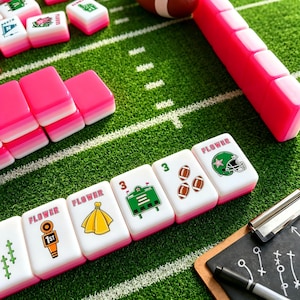 May include: A set of white mahjong tiles with pink backs, featuring football-themed graphics including a green helmet, a scoreboard, a yellow penalty flag, and brown footballs. The tiles are arranged on a green surface resembling a football field.