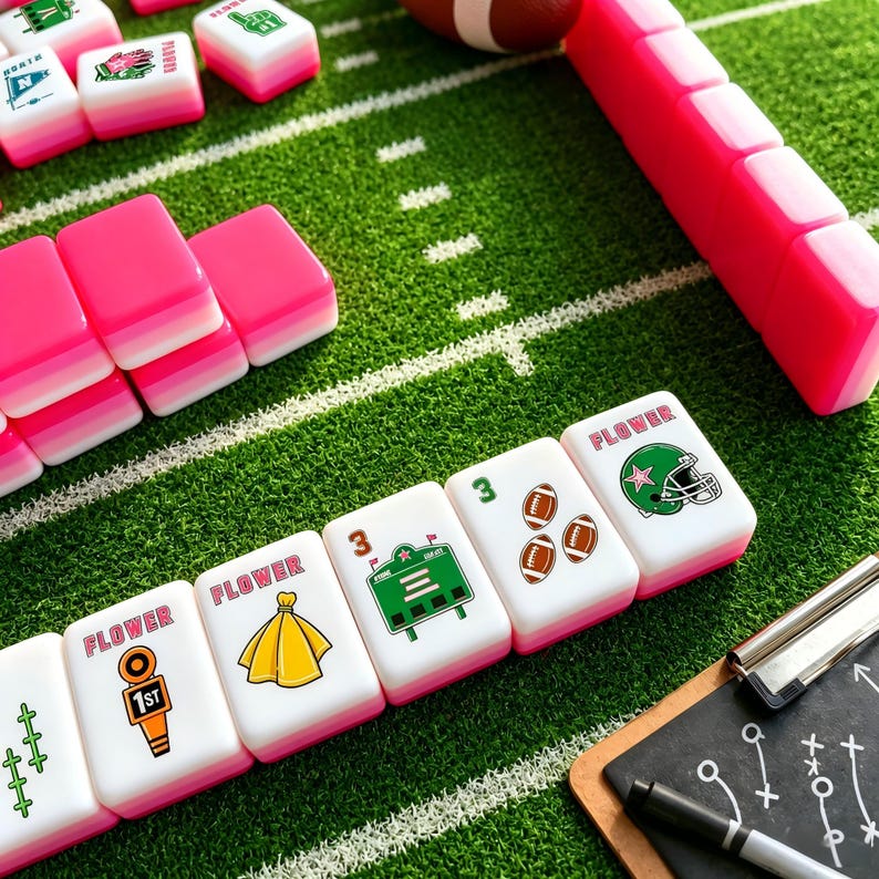 May include: A set of white mahjong tiles with pink backs, featuring football-themed graphics including a green helmet, a scoreboard, a yellow penalty flag, and brown footballs. The tiles are arranged on a green surface resembling a football field.