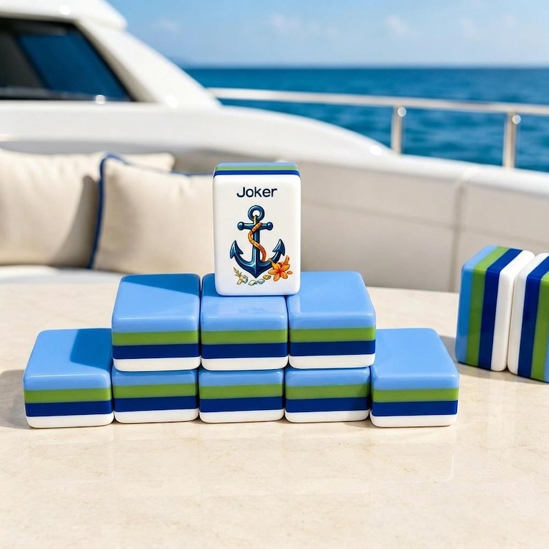 Puede incluir: Un juego de fichas de Mahjong azules y blancas con rayas verdes y blancas. Una ficha es una ficha "Joker" con un dise&ntilde;o de ancla y flor. Las fichas est&aacute;n dispuestas sobre una superficie clara, con un yate al fondo.