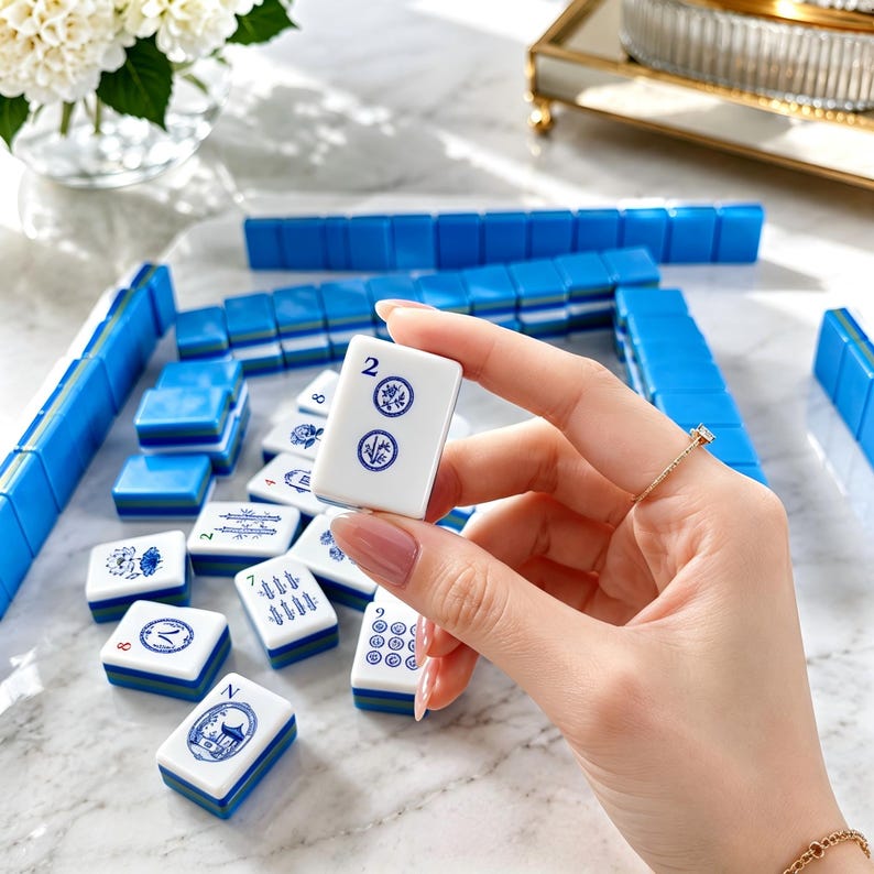 May include: A white mahjong tile with blue markings held in a hand, featuring the number 2 and circular floral designs. The tile has a blue base and is part of a larger set of blue and white mahjong tiles scattered on a white marble surface.