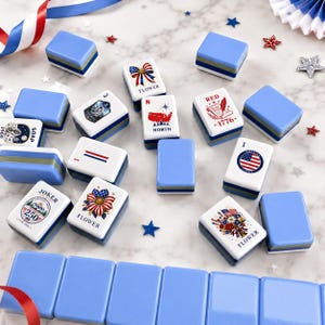 May include: A collection of rectangular mahjong tiles with light blue backs and white faces featuring patriotic American-themed graphics, including flowers, a map, the text 1776, and the word Joker, arranged on a white marble surface with star accents.
