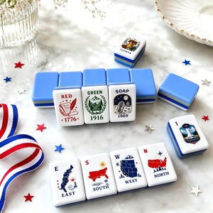 May include: A set of rectangular mahjong tiles with light blue backs and white faces featuring various colorful illustrations and text including RED 1776, GREEN 1916, SOAP 1969, EAST, SOUTH, WEST, NORTH, and a Liberty Bell design labeled FLOWER.
