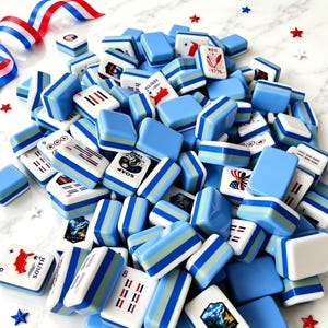 Puede incluir: Un gran mont&oacute;n de fichas de mahjong rectangulares con lados a rayas en azul claro, azul medio y blanco. Las fichas muestran varios gr&aacute;ficos impresos, incluidos s&iacute;mbolos patri&oacute;ticos en rojo, blanco y azul, texto como 1776 y peque&ntilde;os confeti en forma de estrella.