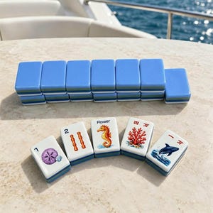 Puede incluir: Un juego de fichas de Mahjong azules y blancas dispuestas sobre una superficie clara. Las fichas presentan varios s&iacute;mbolos y n&uacute;meros, incluyendo un caballito de mar, coral y un delf&iacute;n. Las fichas est&aacute;n apiladas en filas.