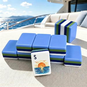 Puede incluir: Un juego de fichas de Mahjong azules y blancas con detalles verdes y blancos. Una ficha presenta un dise&ntilde;o de puesta de sol y la letra "S". Las fichas est&aacute;n dispuestas sobre una superficie clara, con un yate y el mar de fondo.