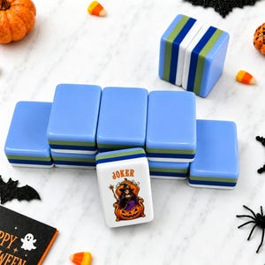 May include: A set of rectangular mahjong tiles with light blue, green, and white layered sides. One tile features a graphic of a witch sitting in a pumpkin with the text JOKER above it. The tiles are arranged in a cluster on a white surface.