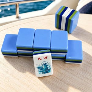 Puede incluir: Un juego de fichas de Mahjong azules con detalles verdes y blancos. Una ficha presenta caracteres japoneses y un dise&ntilde;o de olas. Las fichas est&aacute;n dispuestas sobre una superficie de madera clara, con un vistazo de un fondo de agua azul.