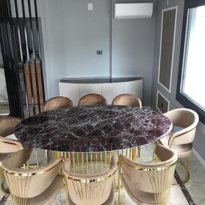 May include: An oval dining table with a dark red marble top and gold metal base. The table is surrounded by beige upholstered chairs with gold accents. The room has a modern design with neutral colors.