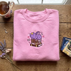 May include: A pale pink sweatshirt with a crew neck. The front displays an embroidered design of a purple dragon wearing a crown, sleeping on a stack of books. The design includes flowers, stars, and a crescent moon.
