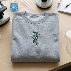 May include: A grey sweatshirt with a crew neck, embroidered with a cartoon DNA strand character. The DNA strand is green, blue, and yellow, with a smiling face and arms raised. The sweatshirt is folded on a wooden surface, with lab equipment visible.