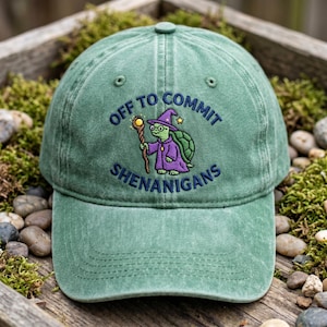 May include: Green baseball cap with the embroidered phrase "OFF TO COMMIT SHENANIGANS." The cap features a cartoon turtle wizard with a staff and purple robe. The hat is made of a soft, worn material.