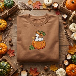 May include: A brown crewneck sweatshirt featuring a central graphic of a white duck sitting inside a large orange pumpkin, with a smaller pumpkin slice resting on the duck's head and two small yellow stars flanking the duck's neck.