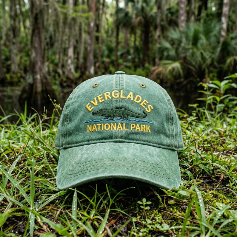 May include: A sage green baseball cap featuring an embroidered design of an alligator above a blue wavy line. The text "EVERGLADES" is arched above the alligator, and "NATIONAL PARK" is embroidered in yellow below the blue line.