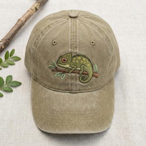 Peut inclure: Casquette de baseball kaki avec un motif brodé d'un caméléon vert sur une branche. Le caméléon a des taches jaunes et une queue enroulée. La casquette a une visière incurvée et un bouton sur le dessus. Couleur neutre.