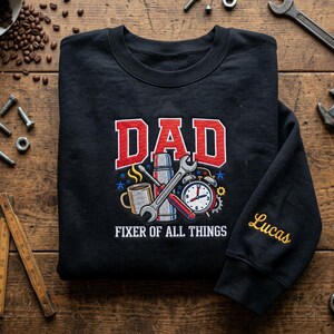 May include: A black crewneck sweatshirt featuring the red text DAD above a graphic of a coffee mug, a thermos, a wrench, and an alarm clock. Below the graphic is the white text FIXER OF ALL THINGS. The name Lucas is embroidered in yellow on the sleeve.