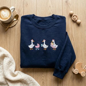 K&ouml;nnte beinhalten: Marineblaues Sweatshirt mit einem gestickten Design von drei G&auml;nsen, jede mit einer rosa Schleife. Eine Gans schiebt einen Kinderwagen, eine andere h&auml;lt eine Kaffeetasse und die dritte tr&auml;gt ein Baby. Eine Tasse Kaffee und Holzspielzeug sind ebenfalls sichtbar.