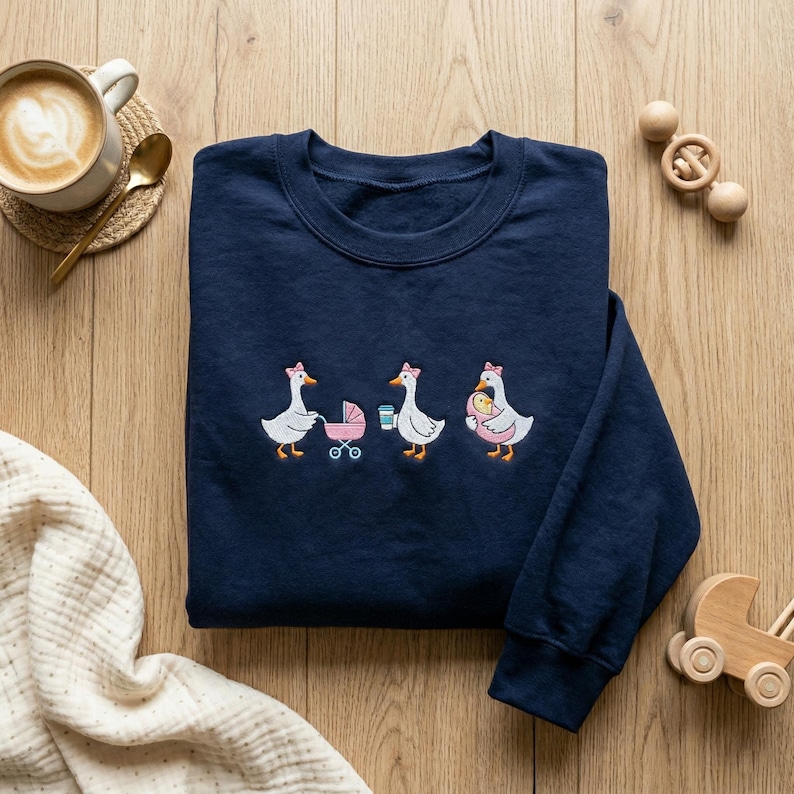 K&ouml;nnte beinhalten: Marineblaues Sweatshirt mit einem gestickten Design von drei G&auml;nsen, jede mit einer rosa Schleife. Eine Gans schiebt einen Kinderwagen, eine andere h&auml;lt eine Kaffeetasse und die dritte tr&auml;gt ein Baby. Eine Tasse Kaffee und Holzspielzeug sind ebenfalls sichtbar.