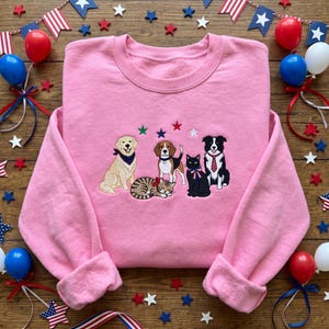 May include: A pink sweatshirt with an embroidered design of a golden retriever, beagle, tabby cat, black cat, and border collie, each wearing accessories. The design is accented with embroidered stars. The sweatshirt has long sleeves and a crew neck.