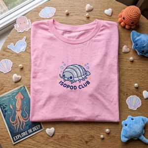 以下が含まれることがあります： 中央に灰色の等脚類が刺繍されたライトピンクのTシャツ。等脚類の周りには小さな青い泡と黄色い星が配置され、その下にはダークブルーの文字で「ISOPOD CLUB」と清潔感のあるサンセリフ体でアーチ状に刺繍されています。