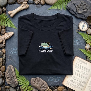 May include: A black short-sleeve t-shirt featuring a graphic of a green creature emerging from water with the text HELLO LAND printed in white below the illustration. The design is centered on the chest of the garment.