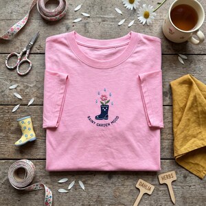 May include: A pink t-shirt with a crew neck, featuring an embroidered design of a rain boot with a flower and the text "Rainy Garden Mood". The shirt is folded on a wooden surface, with gardening accessories and a cup of tea.