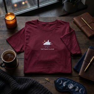 May include: A maroon t-shirt with an embroidered cloud, stars, and heart design, and the text "TINY RAINY CLOUD". The shirt is on a wooden surface with a lit candle, a cup of coffee, and a sleep mask.