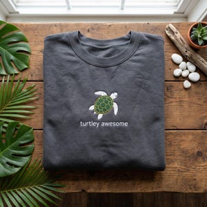 May include: A dark grey crew neck sweatshirt with an embroidered turtle design in white and green. The text "turtley awesome" is below the turtle. The sweatshirt is folded on a wooden surface.