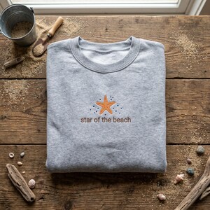 May include: A heather gray sweatshirt with a crew neck, featuring an embroidered orange starfish design and the text "star of the beach" in brown. The sweatshirt is folded on a wooden surface with sand and beach-themed accessories.