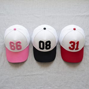 May include: Three baseball caps with white crowns and coloured brims. One has a pink brim with the number 66, another has a black brim with the number 08, and the third has a red brim with the number 31.