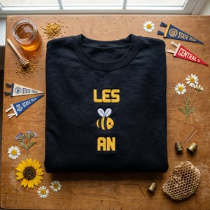 May include: A black crewneck sweatshirt featuring yellow embroidered text that reads LES, followed by a small embroidered bee graphic with white wings and yellow and black stripes, and the yellow embroidered text AN below the bee.
