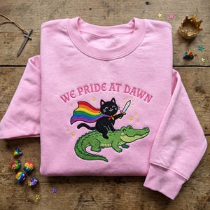 May include: A light pink long-sleeve sweatshirt featuring an embroidered design of a black cat wearing a rainbow cape and holding a sword while riding a green alligator. The text WE PRIDE AT DAWN is embroidered in pink above the cat and alligator.