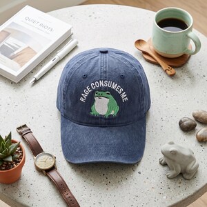 May include: A navy blue baseball cap featuring an embroidered green frog graphic centered on the front. Above the frog, the text "RAGE CONSUMES ME" is stitched in white capital letters. The cap has a structured crown and a curved brim.