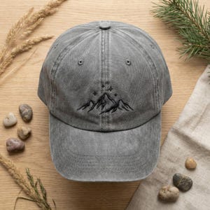 May include: A charcoal grey baseball cap with a distressed, washed-out finish. The front panel features a black embroidered line art design of a mountain range topped with three small four-pointed stars. The cap has a curved brim and stitching.