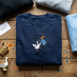 May include: Navy blue sweatshirt with an embroidered design of a white goose spilling paint from a blue paint can. The text "GOT TO SILLY" is above the goose. Additional sweatshirts are visible in the background.