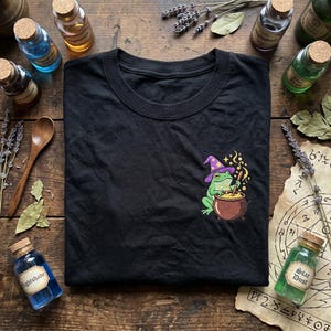 May include: Black t-shirt featuring an embroidered design of a green frog wearing a purple wizard hat, stirring a cauldron. The shirt is surrounded by apothecary bottles, herbs, and a wooden spoon, creating a mystical theme.