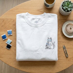 Peut inclure: Sweat-shirt blanc à col rond avec une broderie d'un chat utilisant un microscope. Le chat porte des lunettes de protection. Les détails sont bleus et noirs. Le sweat-shirt est posé sur une table en bois.