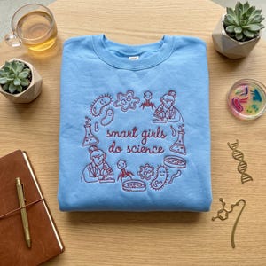 Peut inclure: Un sweat-shirt bleu clair avec la phrase brodée "smart girls do science" entourée d'illustrations scientifiques rouges. Le sweat-shirt est présenté sur une surface en bois avec des éléments décoratifs.
