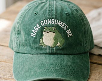 May include: A forest green baseball cap featuring an embroidered illustration of a frog with a cream-colored belly. The text RAGE CONSUMES ME is embroidered in white capital letters in an arc above the frog on the front of the cap.