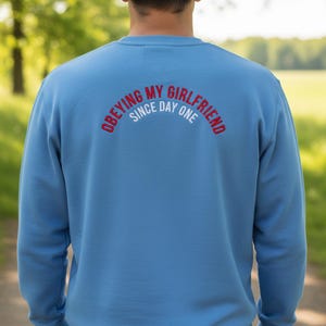 May include: A light blue sweatshirt with the text "OBEYING MY GIRLFRIEND SINCE DAY ONE" in red and white arched across the back. The sweatshirt is a solid color and has a crew neck.