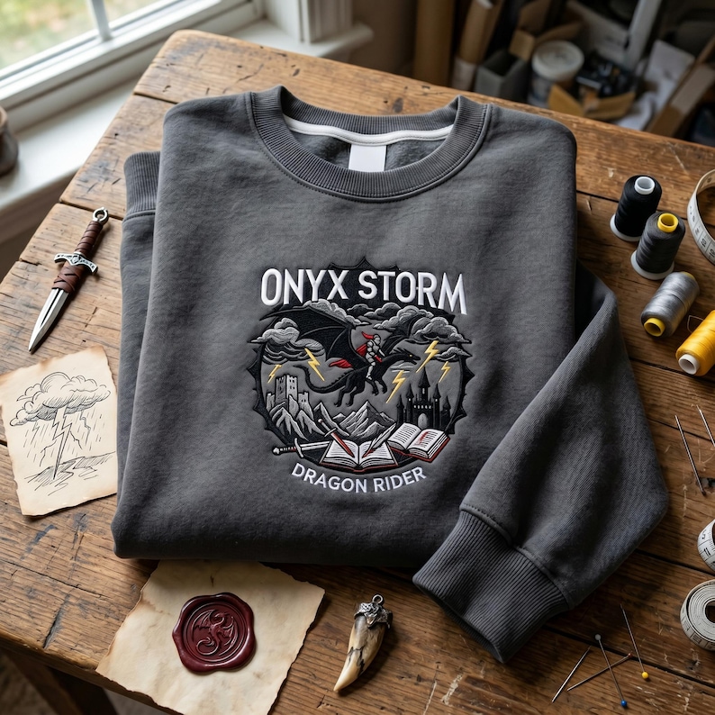 May include: A dark gray sweatshirt with the text "ONYX STORM" above a graphic of a dragon rider. The graphic includes a dragon, castle, and open books. The sweatshirt is folded on a wooden surface with other fantasy-themed items.